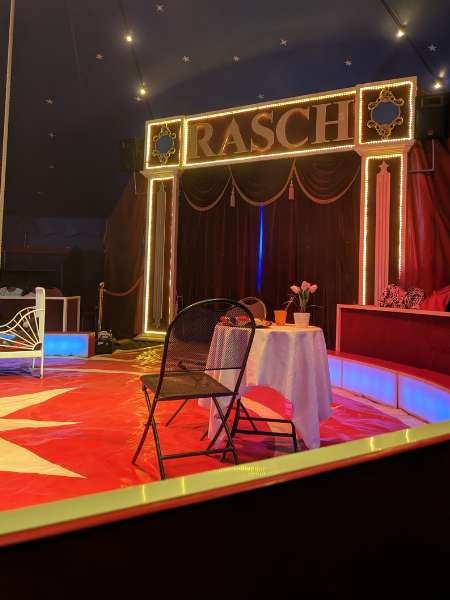 Ausschnitt aus einer Zirkusmanage, darin steht ein gedeckter Tisch und ein Stuhl. Im Hintergrund sieht man den Namen des Zirkus (Rasch) über dem Eingang für die Artisten.