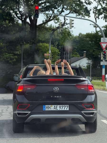 Schwarzes Cabrio von hinten steht vor einer roten Ampel. Die vier Insassen strecken die Hände nach oben.