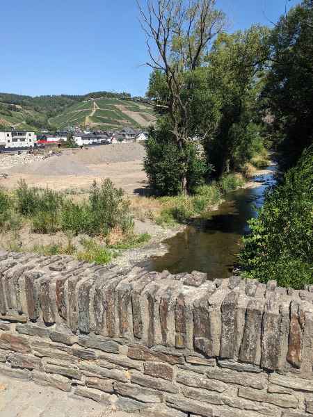 Steinbrücke über einem kleinen Fluss, im Hintergrund Berge und Häuser.